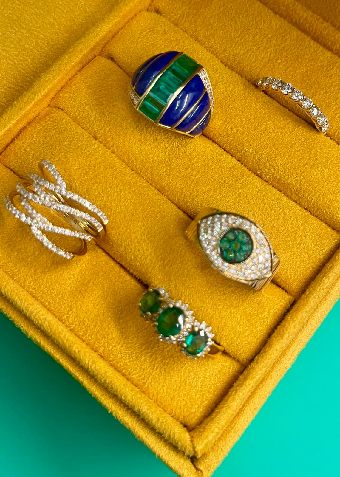 Anéis com esmeraldas, lápis-lazúli e diamantes em estojo de camurça mostarda sobre fundo verde-água